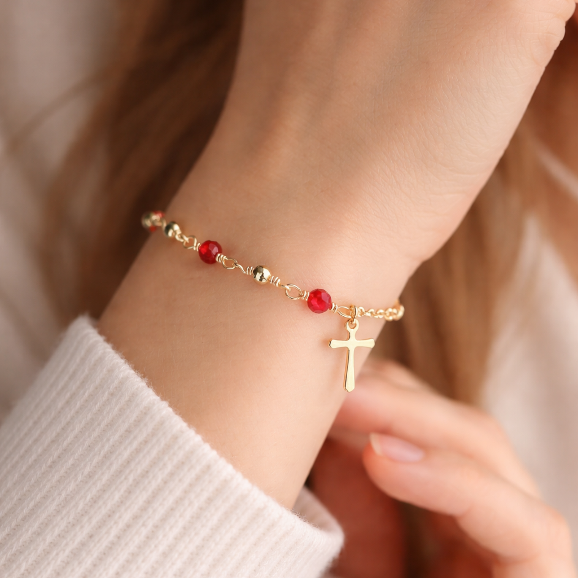 Gold bracelet with red beads and a cross charm on a wrist.