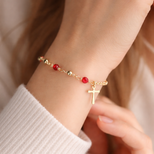 Gold bracelet with red beads and a cross charm on a wrist.