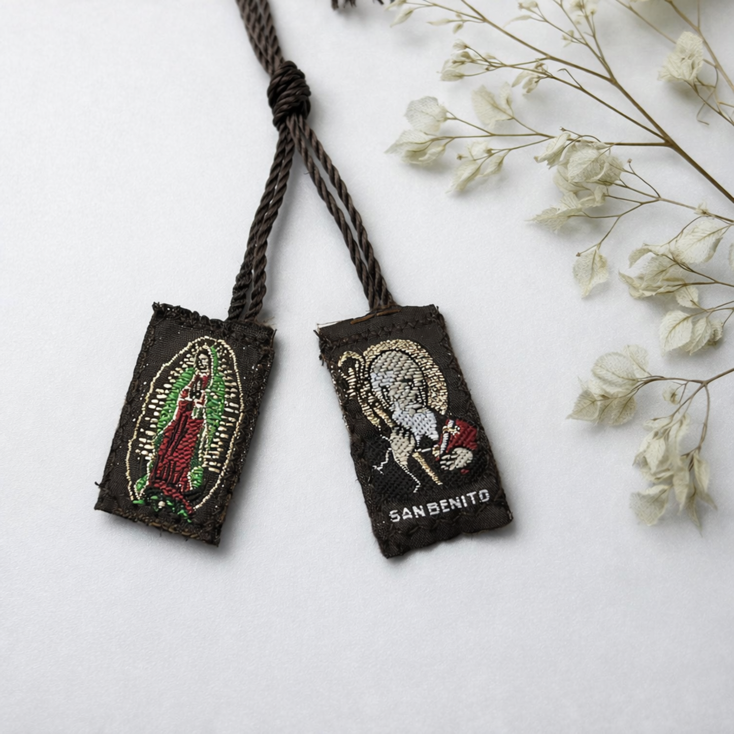 Two embroidered pouches with Virgin Mary and Saint Benedict designs on a white surface with dried flowers.