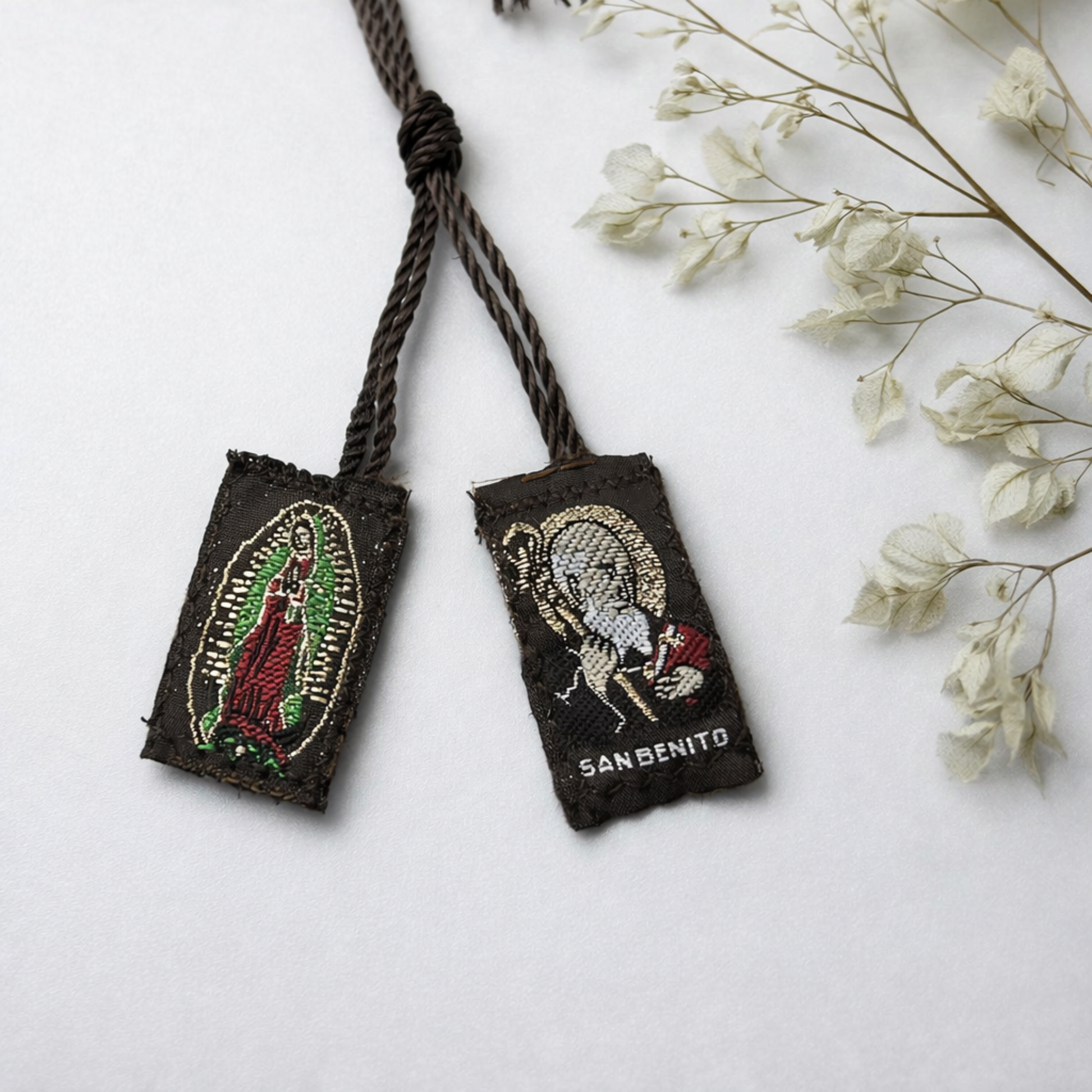 Two embroidered pouches with Virgin Mary and Saint Benedict designs on a white surface with dried flowers.