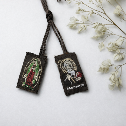 Two embroidered pouches with Virgin Mary and Saint Benedict designs on a white surface with dried flowers.