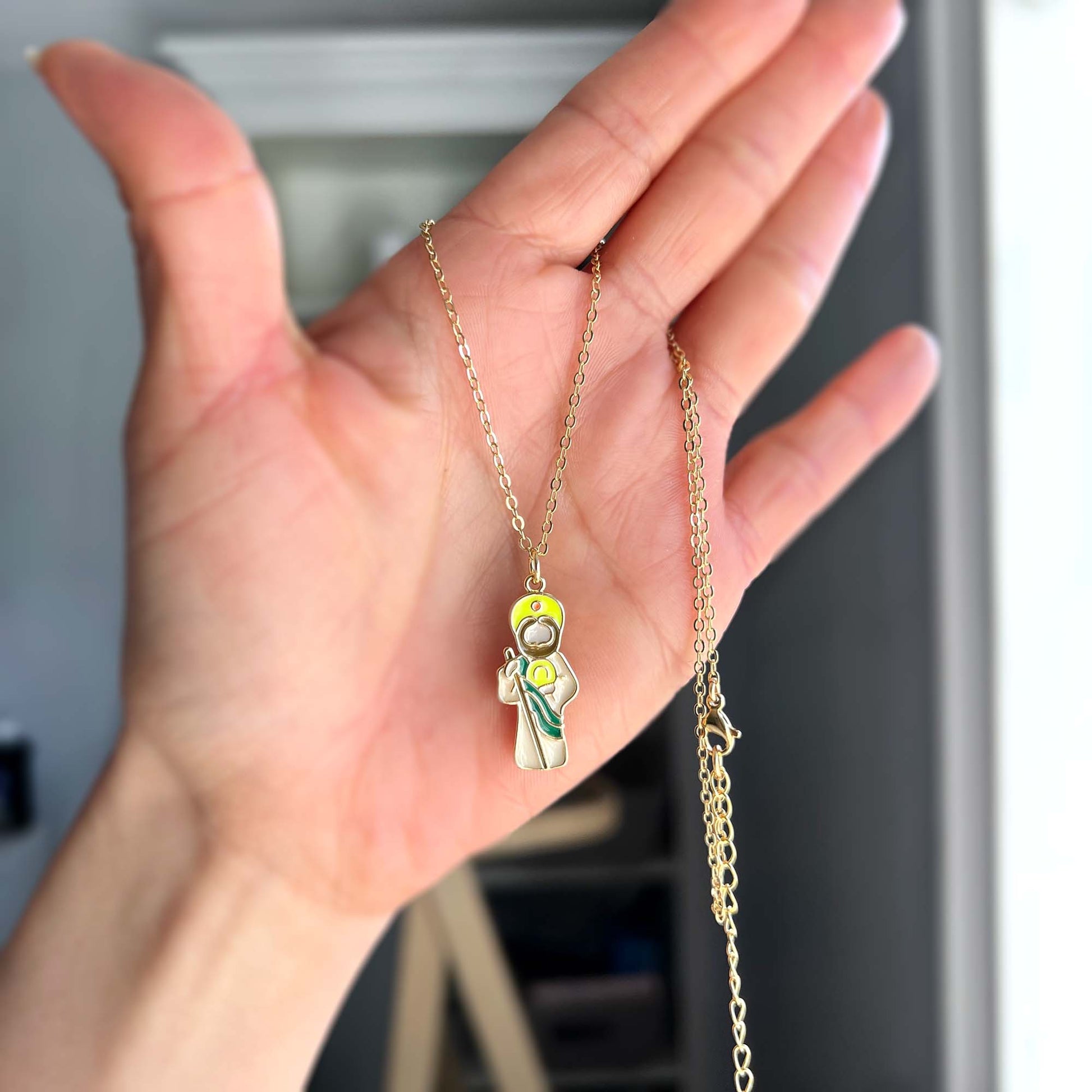 Hand holding a gold necklace with a colorful pendant of Saint Jude against a blurred background