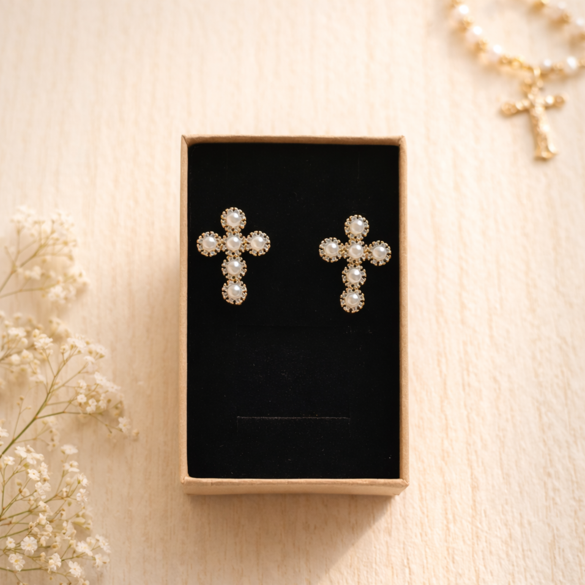 Cross-shaped earrings with pearl details in a black box on a beige surface with flowers.