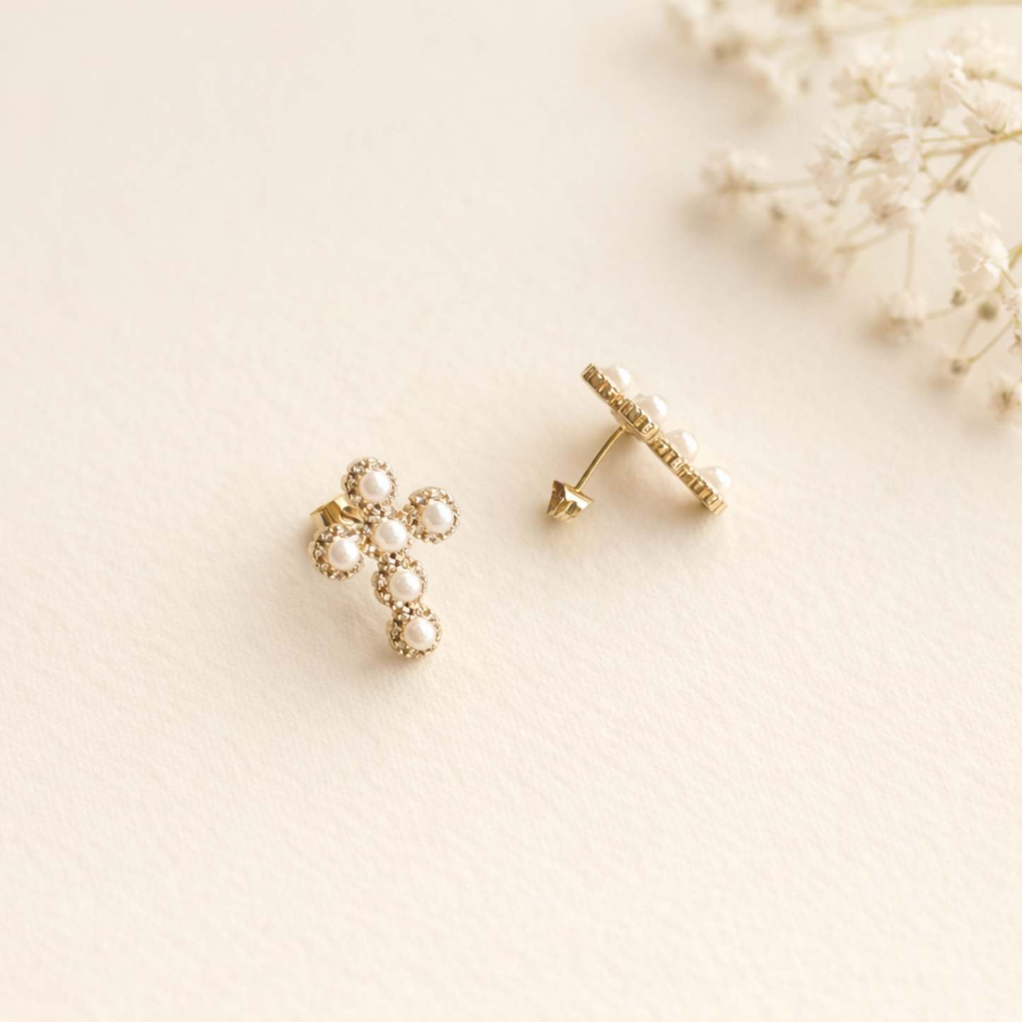 Gold cross-shaped earrings on a light beige background with dried flowers.