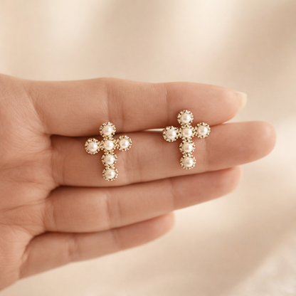 Gold cross earrings with pearl-like stones held in a hand against a neutral background