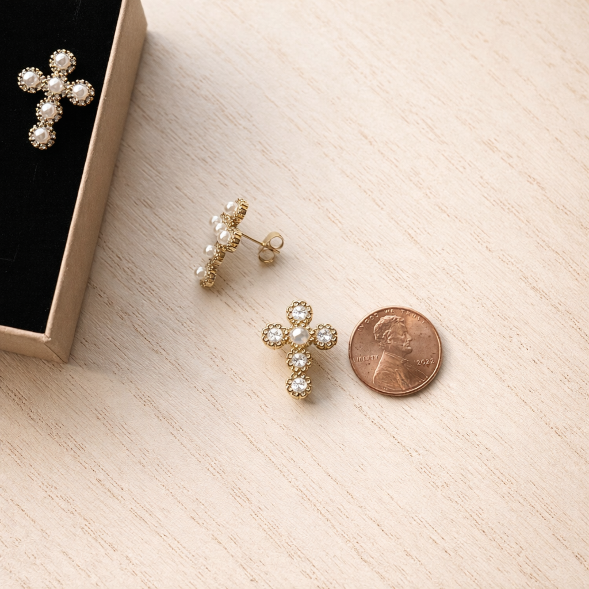 Gold cross-shaped earrings with pearl details on a light wooden surface next to a penny for scale.