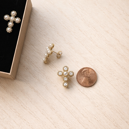 Gold cross-shaped earrings with pearl details on a light wooden surface next to a penny for scale.