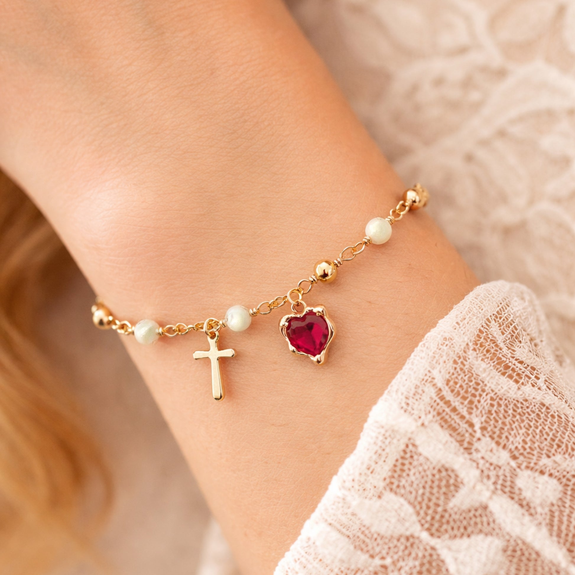 Gold bracelet with heart charm, cross charm, and pearl beads on a wrist.